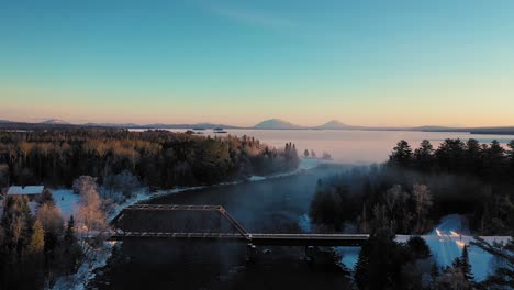 flying over the mist of a curving winter river past a railroad trestle towards a snow covered frosted lake with two prominent mountains in the distance