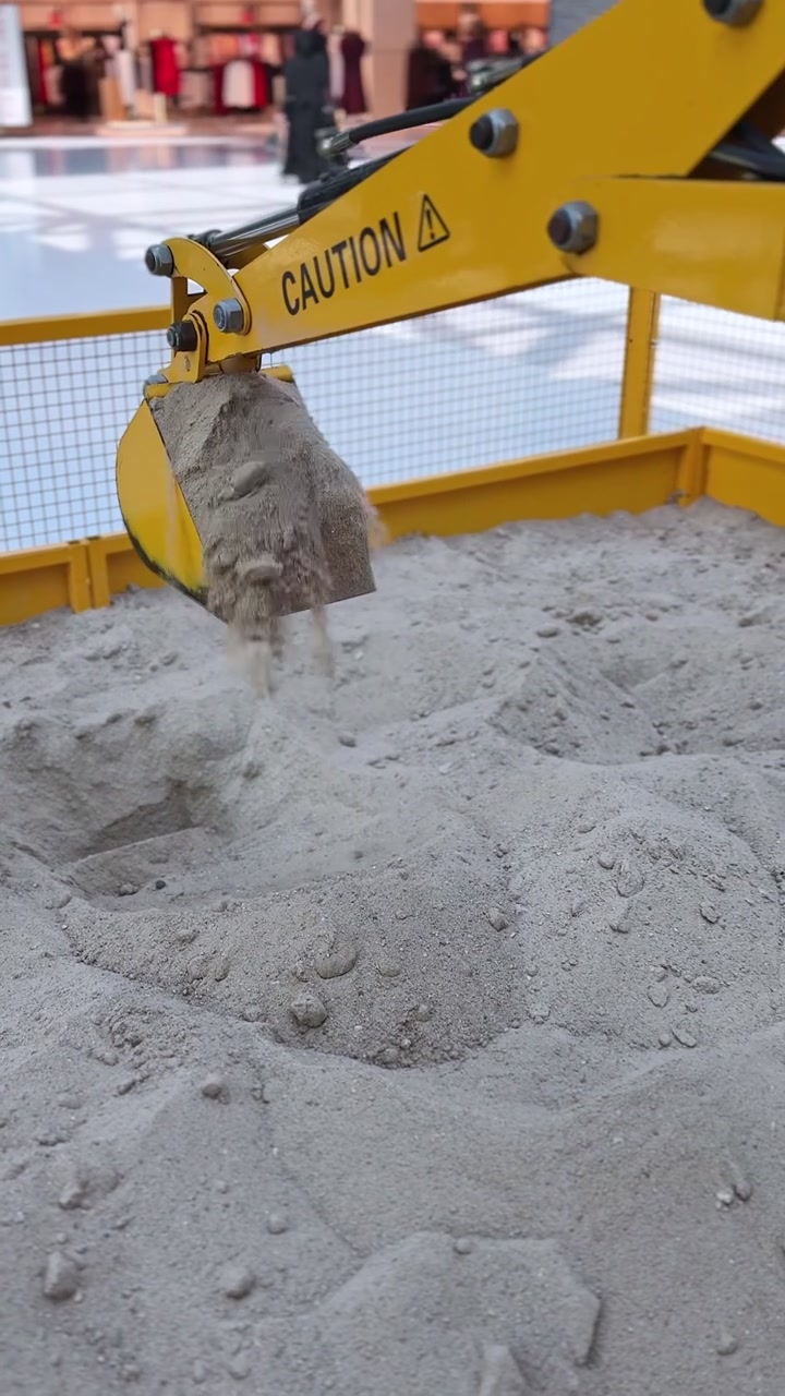 Et nærbillede i vertikalt format af en gul miniature legetøjsgraverspand, der graver og skovler løst sand. Den mekaniske arm opererer inde i et indhegnet indendørs sandkasseområde i et lyst oplyst indkøbscenter.