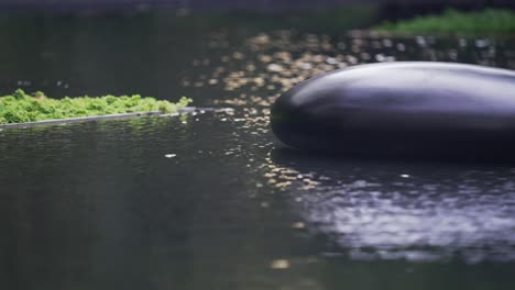 Ein-Großer-Schwarzer-Stein-Im-Wasser,-Mit-Einer-Reflektierenden-Wasseroberfläche,-Die-Eine-Abstrakte-Und-Heitere-Note-Verleiht