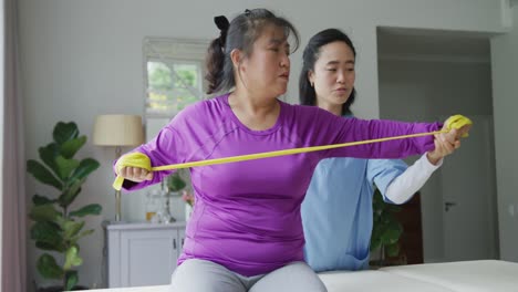 Asian-female-physiotherapist-helping-female-patient-exercise-arms-with-exercise-band-at-surgery