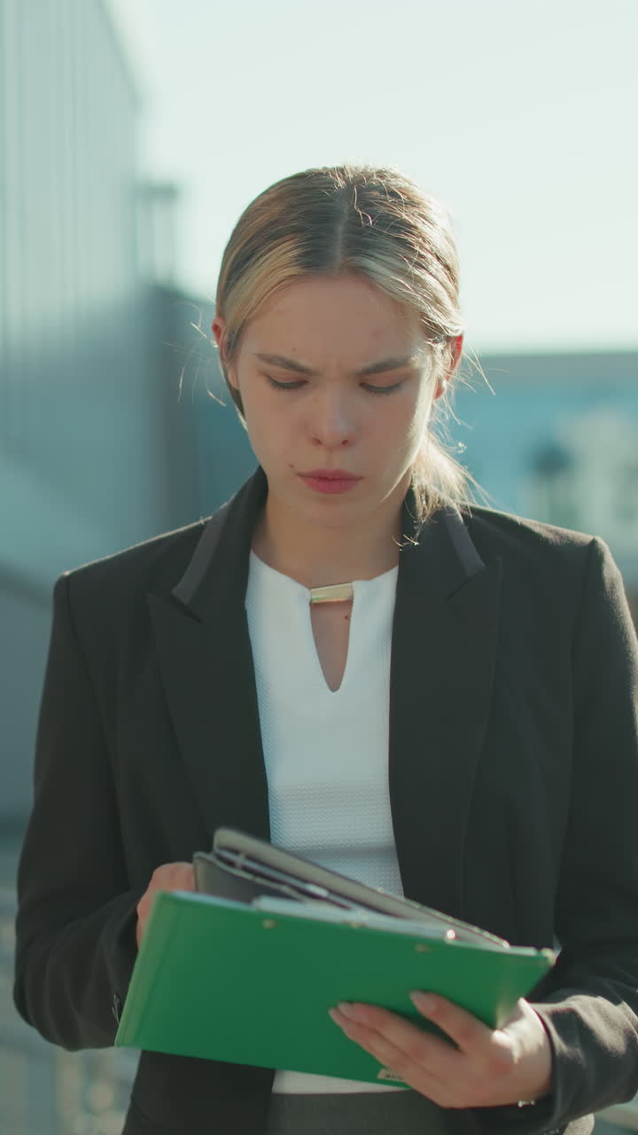 Focused female CEO outdoors in formal attire reviewing documents in folder while walking near modern glass building with pedestrians and parked cars in background on bright sunny day