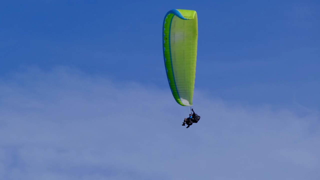 Bottom view of tandem paraglider with green and yellow parachute flying circes on a bright blue sunny day