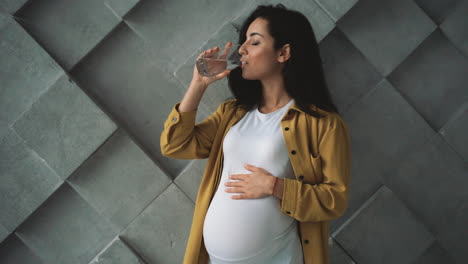 Young-happy-pregnant-woman-holding-and-stroking-her-belly,-drinking-water