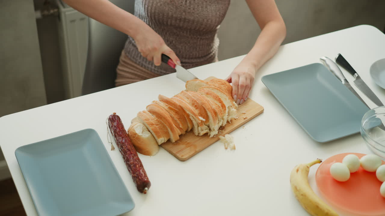 High angle hand view of culinary artist cutting loaf of bread with knife on white table next to eggs banana and bowls in kitchen under bright sunny morning light streaming through window behind
