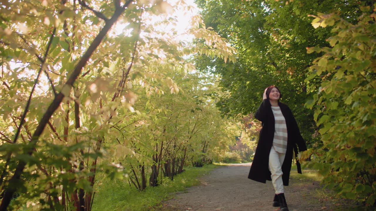 Joyful walker with headphones holds autumn leaf, listens to music and spins beneath sunlit arboretum canopy, relaxed stroll along leafy path, playful turn, serene mood, casual coat boots style