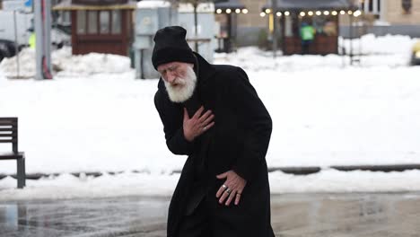 an elderly man clutches his chest while walking through a snowy scene, dressed in dark winter clothing.