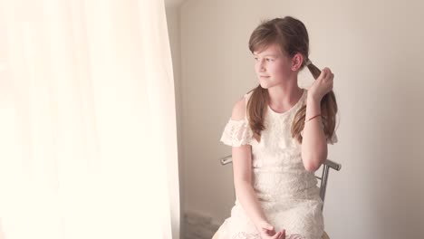 Child-Portrait-of-Girl-Sitting-in-Chair-By-Window-on-Daylight-in-White-Dress-Moving-Her-Pigtals-to-Look-Perfetcly-and-Looks-Through-Window