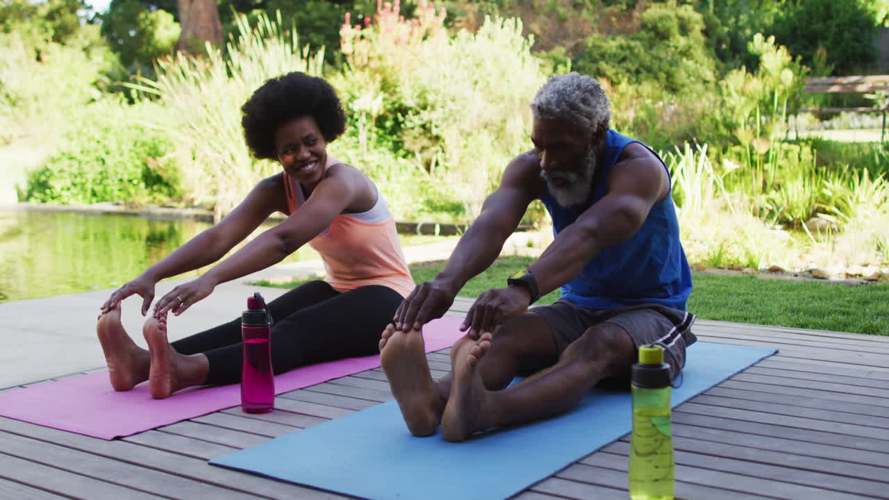 una pareja de ancianos afroamericanos haciendo ejercicio al aire libre sentados estirándose en un jardín soleado