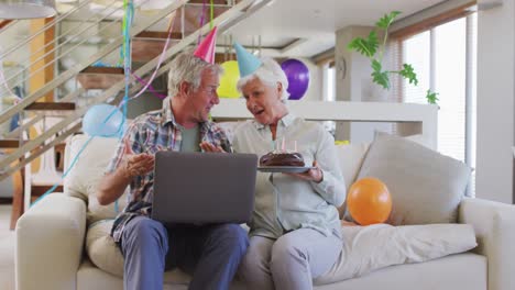 Pareja-Caucásica-Mayor-Sosteniendo-Pastel-Y-Celebrando-Cumpleaños-Teniendo-Un-Video-Chat-En-Una-Computadora-Portátil-En-Casa