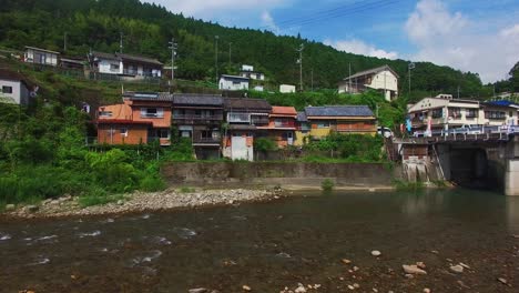 drone view travels forward above a gently flowing river, unveiling charming residences nestled beside its waters in a japanese village