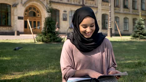 young muslim girl in hijab is sitting on lawn and reading magazine, watching at camera, smiling, builging on background, religious concept, relax concept