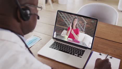 médico afroamericano que usa auriculares de teléfono tomando notas mientras hace una videollamada en una computadora portátil