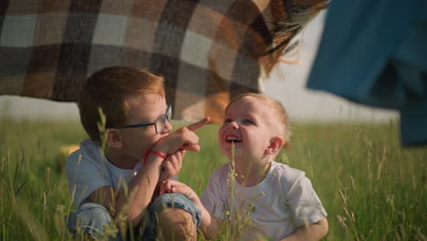 a woman in a blue gown, her face not visible, holds a plaid scarf as the wind gently blows it in a grassy field. beneath the scarf, two happy children in white shirts point at their surroundings
