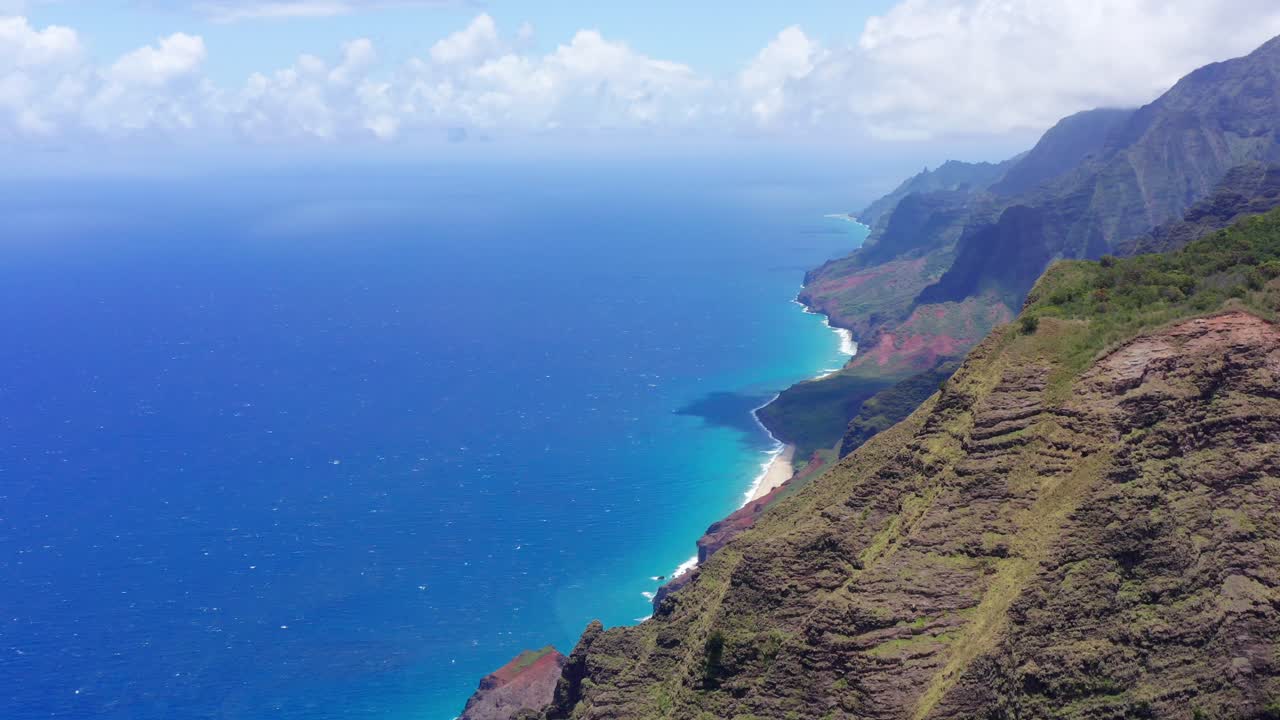 4K drone shot, tilting down of a ridge on the Na Pali Coast on the island of Kauai. It's a beautiful sunny day with puffy white clouds, crystal blue and turquoise water, rocky mountain ridge