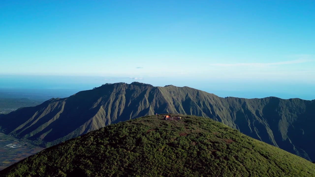Tent campsite on a grassy mountain summit above rugged volcanic landscapes beneath clear blue skies in Lombok Indonesia, cinematic orbit drone footage