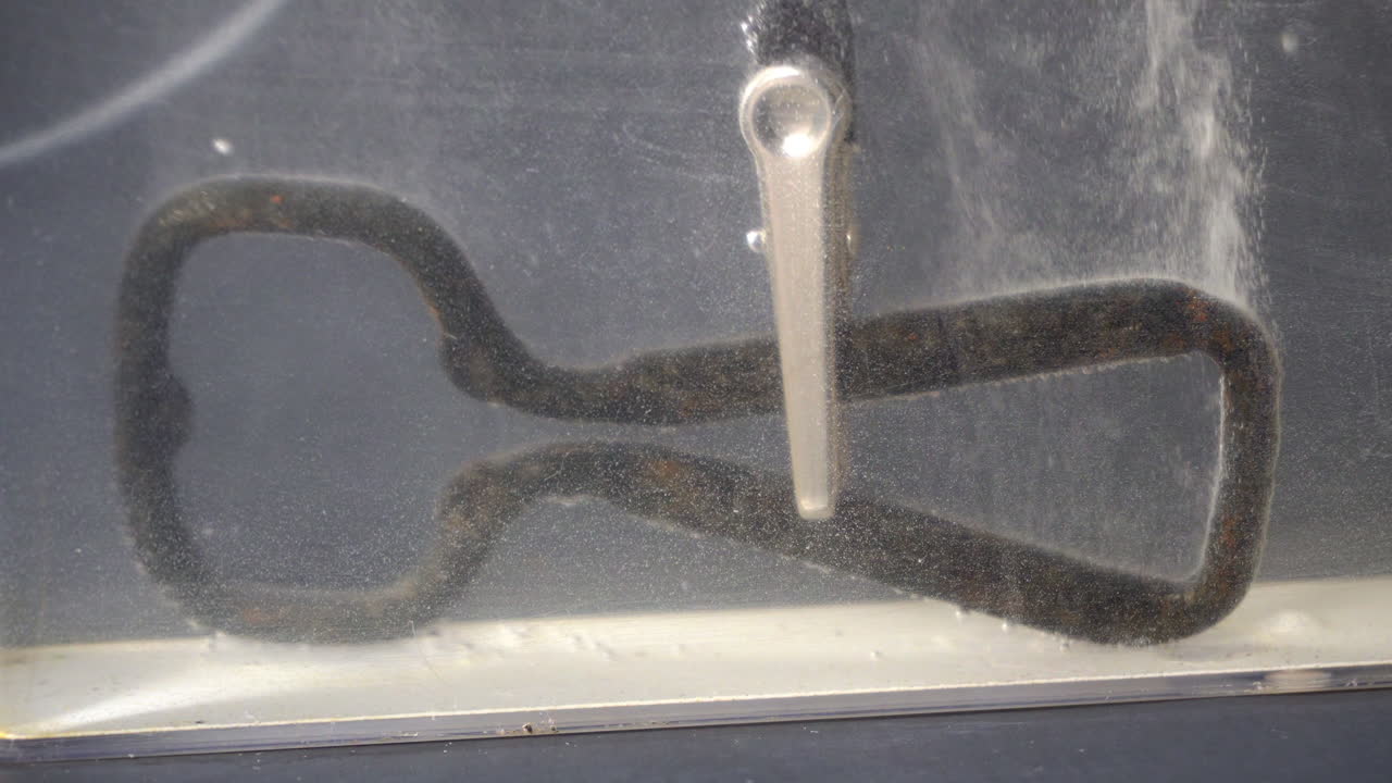 Rusty bottle opener releases bubbles during electrolytic cleaning. Patches of rust are replaced with new grey metal