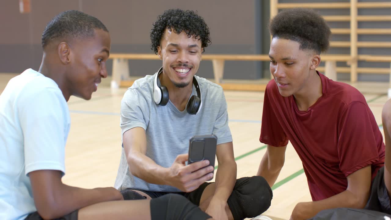 Diverse male friends wearing knee pads on gym floor tilting smartphone, sharing clip and laughing