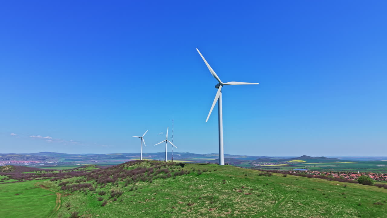 Turbine eoliche su una collina in un paesaggio verde di giorno con cielo azzurro e montagne in lontananza.
