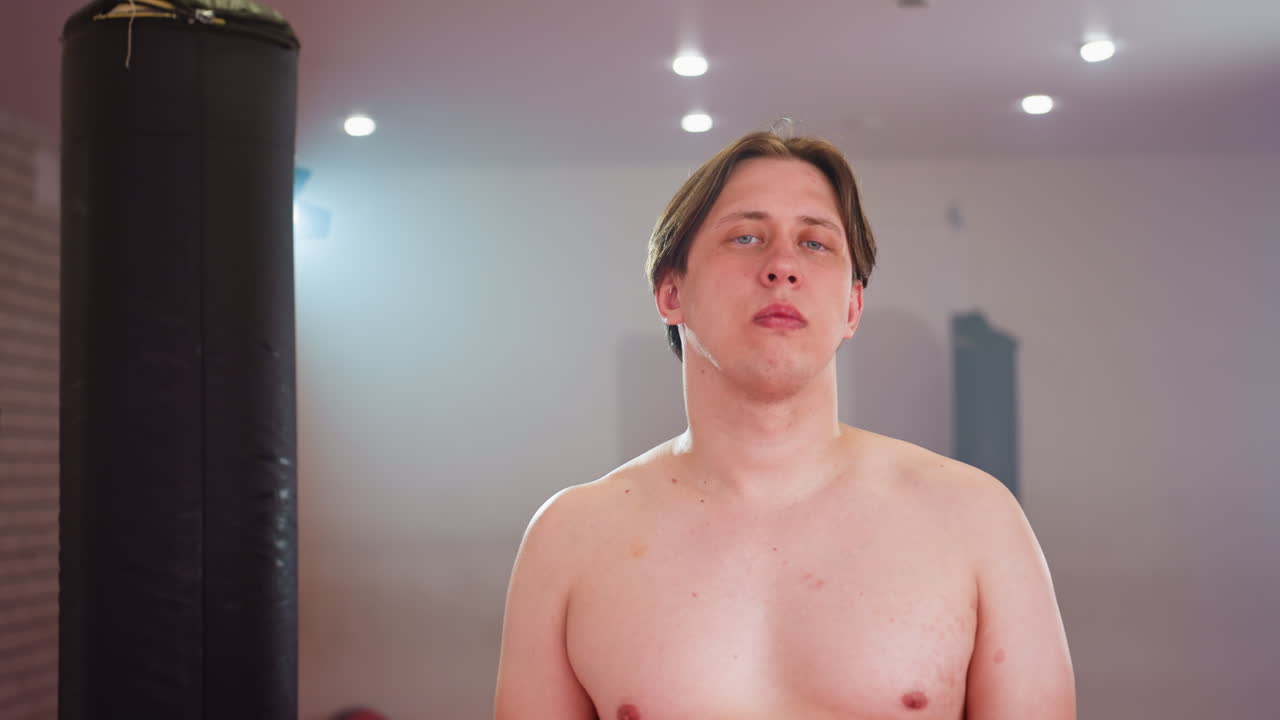 Fighter stands shirtless in gym environment with punching bags and training equipment, body exposed under strong lighting, representing preparation, discipline with determined expression and focus