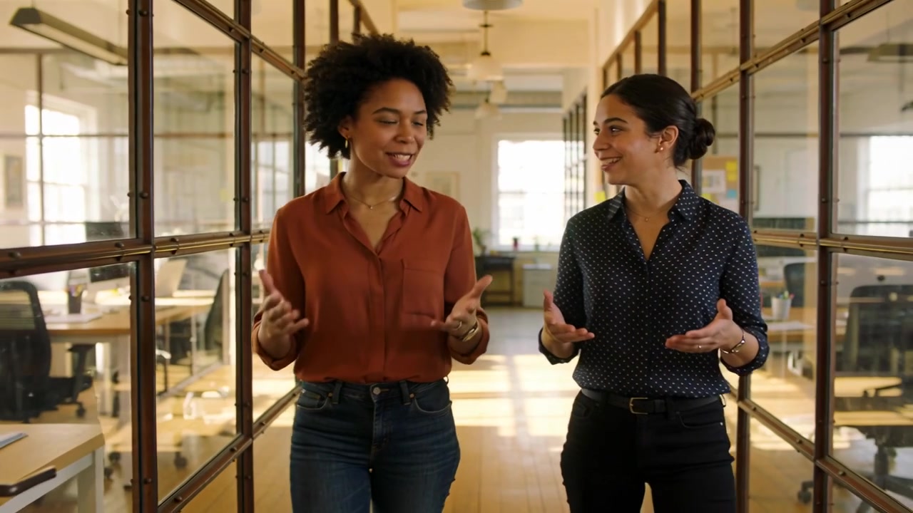 Colleghi che camminano e discutono di lavoro in una sala di vetro; a sinistra, top color ruggine e jeans, a destra, top stampato e pantaloni neri.