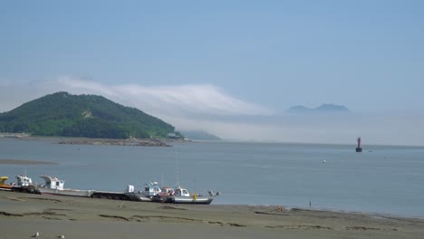 Fisher's-boats-stuck-in-the-mud-on-low-tide-at-Ganghwado-island-in-South-Korea,-haze-and-vapor-over-mountain-peaks,-Red-navigational-buoy-floating-in-Yellow-sea-between-islands