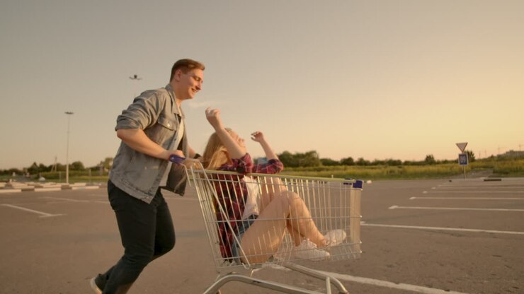Cheerful people a couple a man and a woman at sunset ride on supermarket trolleys in slow motion.