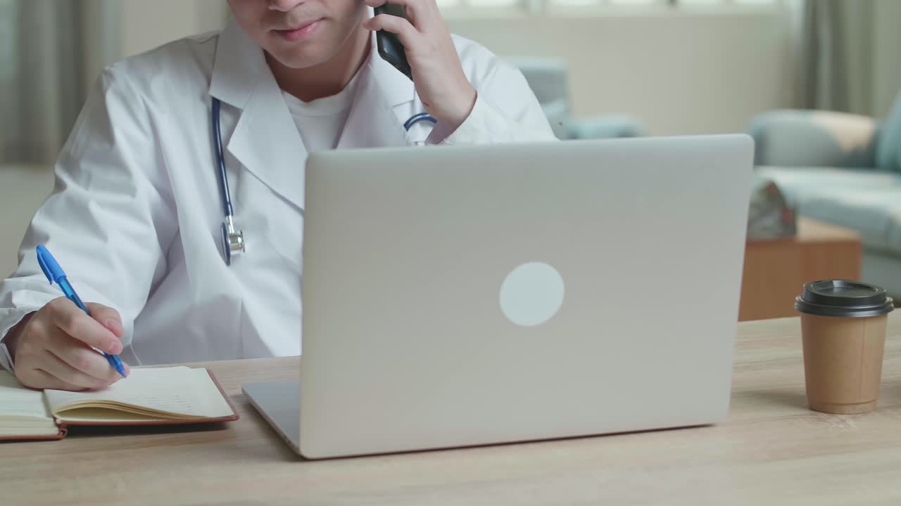 Male Doctor Talking On Phone And Writing On Notebook At Home Office, Video In 4K