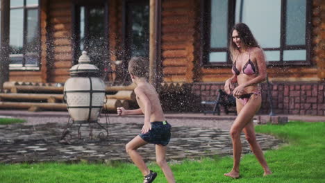 mother and father playing with children on the lawn of the house pouring water from a hose