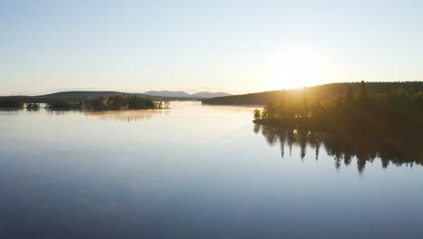 Aerial-flight-over-a-foggy-lake-during-sunrise-with-islands-with-green-trees-and-mountains-in-the-background-filmed-in-Lapland-Finland