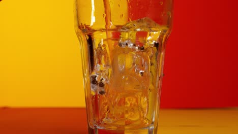 Static-shot-of-a-filled-glass-with-clear-ice-cubes-while-orange-juice-poured-in-on-yellow-red-background