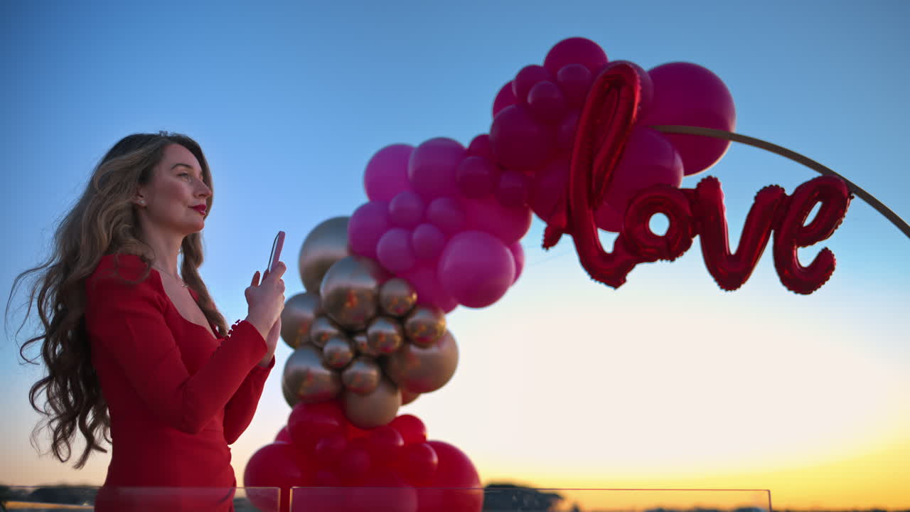 Beautiful young woman in red dress taking photos of the sunset near the red and pink balloon arch spelling out the word "love"