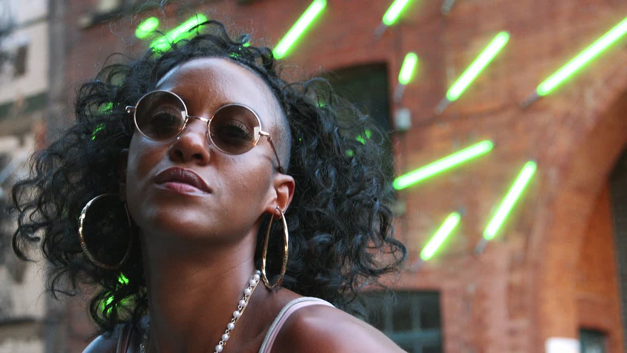 Fashionable young black woman wearing round sunglasses posing to camera in an urban street, close up, low angle, blurred neon strip lights in background