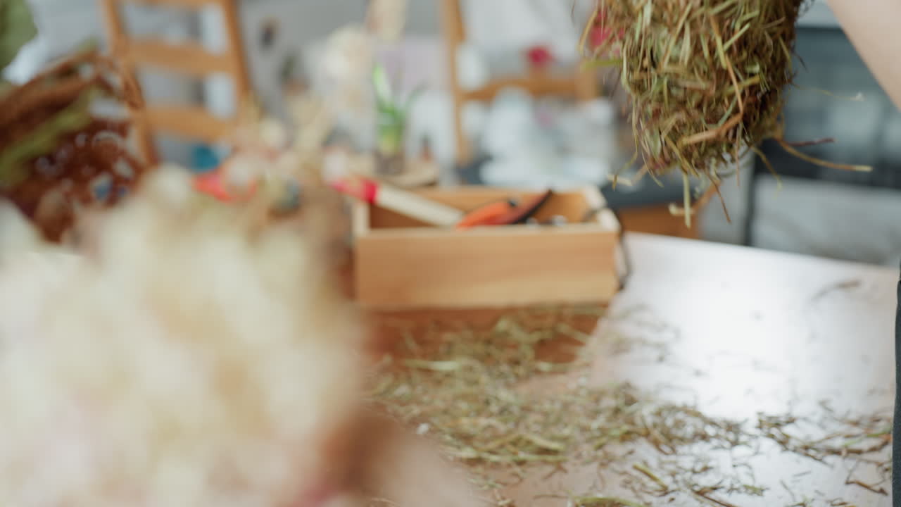 Hands with red nails holding unfinished straw wreath, preparing natural decoration with hay on table surrounded by craft supplies, moment of artisan process of handmade eco friendly rustic ornament