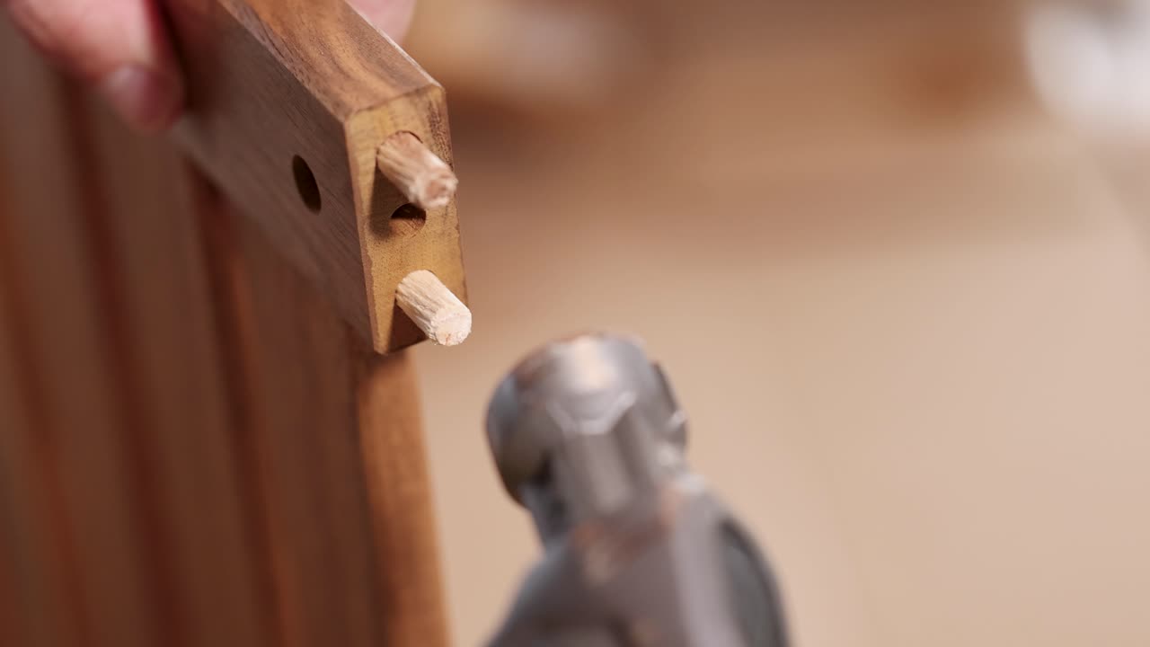 Hands insert a dowel into wood, securing it with a hammer. Close-up view highlights precision and craftsmanship