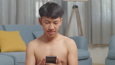 close up of asian teenager boy sitting and using smartphone after doing the shirtless workout at home