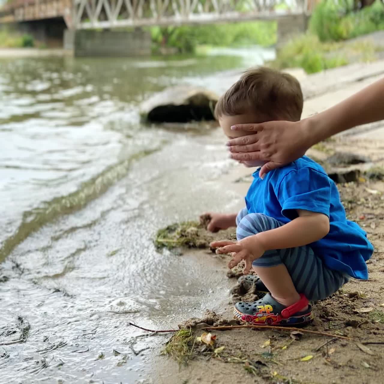 Caucasian kid sits squatted on the shore close to the water. Baby boy picks and throws back wet sand