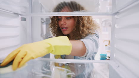 view looking out from inside empty refrigerator as woman wearing rubber gloves cleans shelves
