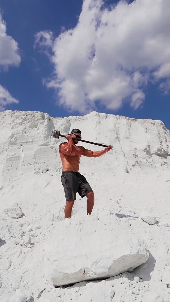 Muscular man on the background of white mountain. Shirtless guy with iron hammer on the natural background of large mountain in summer. Vertical video