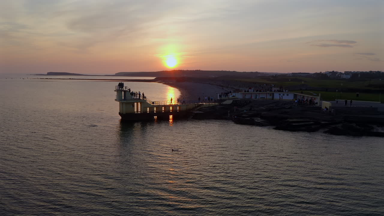 Super slow motion of sun setting behind diving tower with soft ocean reflections with people gathered on railing