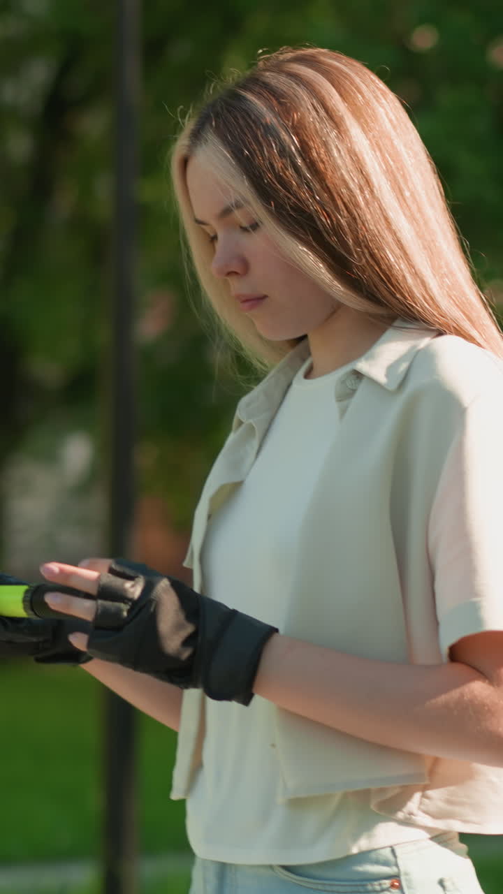 vista lateral de una mujer joven caminando al aire libre, examinando la bomba de aire verde con una expresión enfocada y reflexiva, la luz del sol se refleja en su cara, con un fondo borroso de árboles y edificios distantes