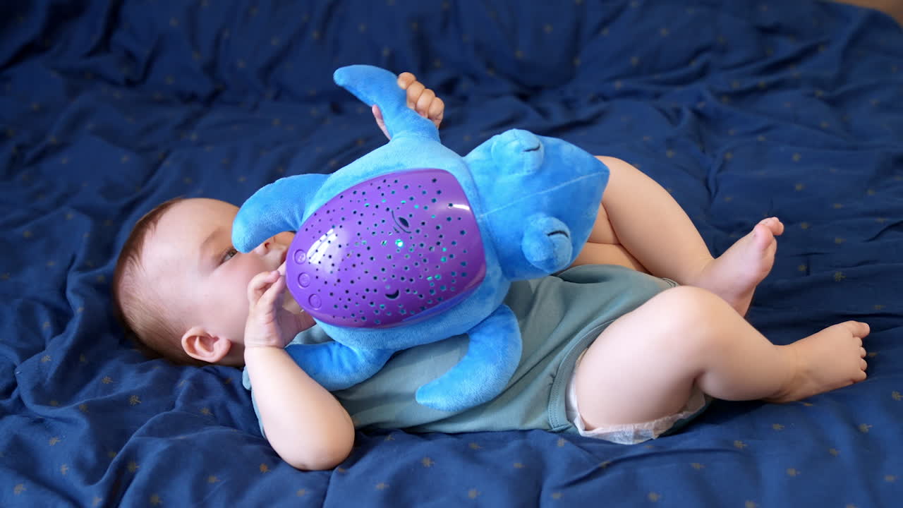 Little infant baby boy lies holding a toy. Healthy child playing with big blue soft turtle. Blue backdrop.