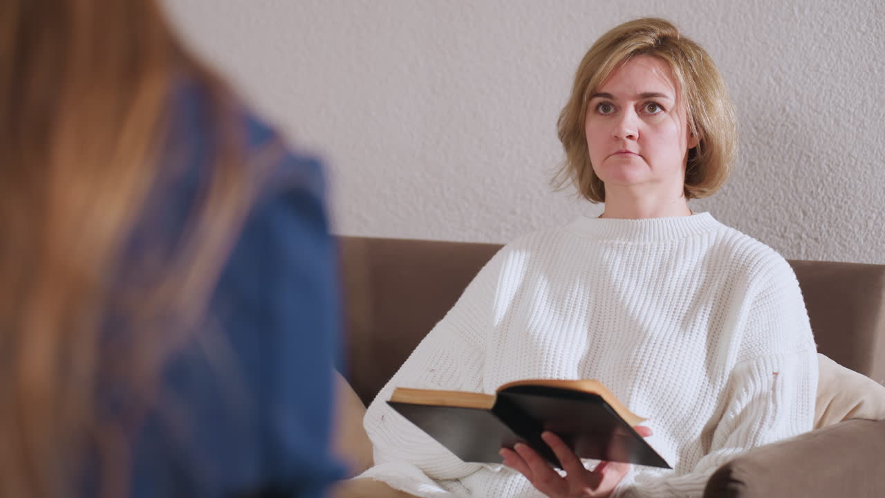Healing seeker seated on sofa opening book while lifting head to listen to therapist during thoughtful moment in calm therapy room, highlighting emotional engagement and introspective exploration