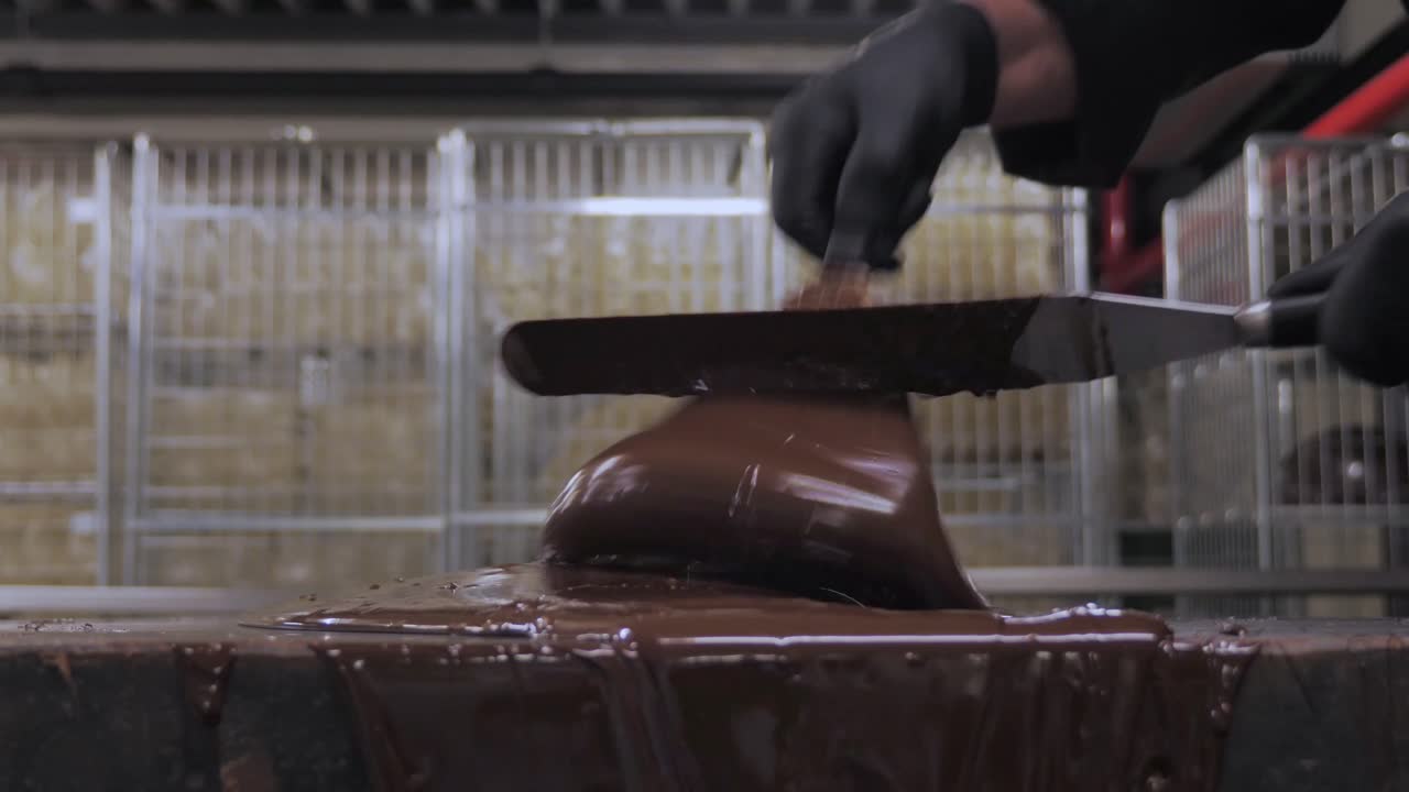 Primo piano di un lavoratore con i guanti che raschia cioccolato fondente fuso denso da un tavolo in un contenitore