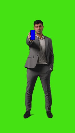 Vertical-Video-Full-Length-Shot-Of-Businessman-Holding-Blue-Screen-Mobile-Phone-Towards-Camera-Standing-Against-Green-Screen