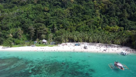 Outrigger-Tour-Boats-approaching-Seven-Commandos-Beach-of-El-nido-with-Tourists-swimming-in-turquoise-clear-water-with-tropical-lush-green-background