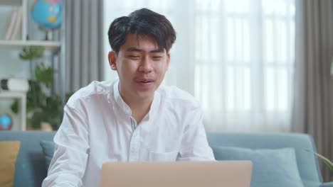 smiling asian man having video call on laptop computer while sitting on sofa in the living room