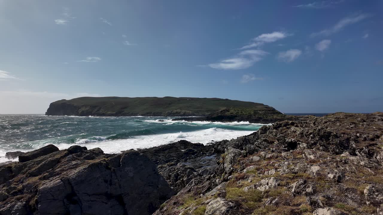Windswept coast with jagged rocks, crashing waves, and a distant island under a bright blue sky