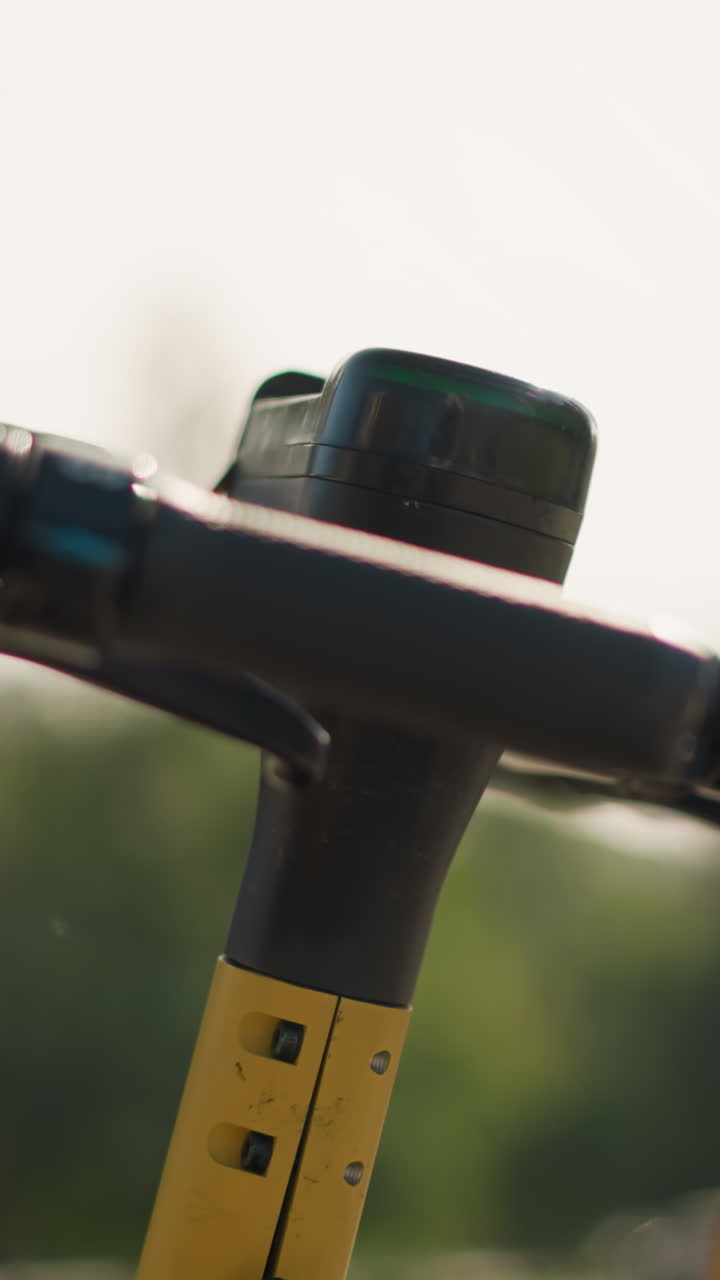 Primer plano del manillar de un patinete eléctrico con barra amarilla y bokeh suave, estacionado en el muelle de la ciudad con una luz cálida de la mañana. La escena tranquila evoca la rutina lenta de un viajero, estudiante, mensajero y turista.
