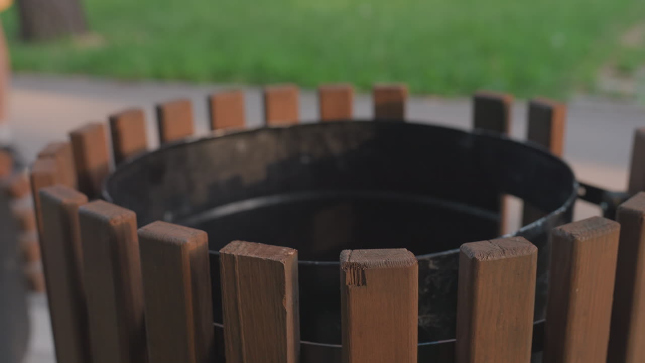 Park Jogger Walks Past Bin, Athletic Figure In Yellow Shorts Running Along Sidewalk With Wooden Waste Container Rim In Foreground, Empty Interior Exposed And Green Lawn Backdrop, Moment Of Public