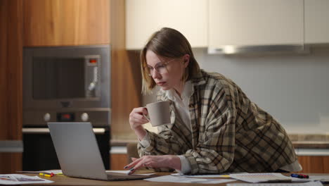 Eine-Frau-Trinkt-Kaffee-Und-Schaut-Auf-Ihren-Laptop,-Während-Sie-Die-Dokumente-Betrachtet